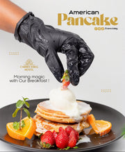 A close-up of a hand wearing a black glove placing a strawberry on top of a plated stack of pancakes with whipped cream, strawberries, orange slices, and mint, with the text 'American Pancake' and 'Carro Edeg' on a light background.