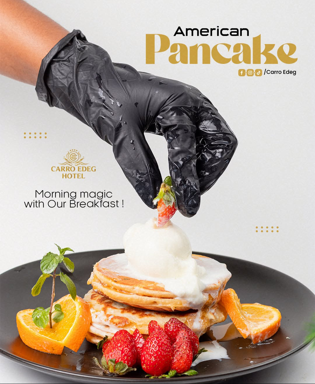 A close-up of a hand wearing a black glove placing a strawberry on top of a plated stack of pancakes with whipped cream, strawberries, orange slices, and mint, with the text 'American Pancake' and 'Carro Edeg' on a light background.