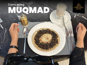 Top-down view of a table setting at Carro Edeg Hotel, featuring a plate of Muqmad (a traditional Ethiopian injera flatbread stuffed with stew) on a white plate, alongside a glass of coffee, a small gold bottle, and a person's hands on a dark marble table.