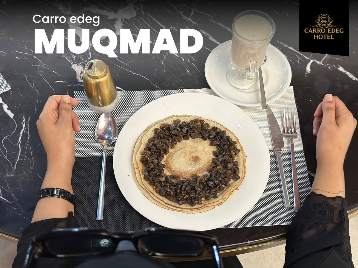 Top-down view of a table setting at Carro Edeg Hotel, featuring a plate of Muqmad (a traditional Ethiopian injera flatbread stuffed with stew) on a white plate, alongside a glass of coffee, a small gold bottle, and a person's hands on a dark marble table.