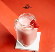 A glass mason jar filled with a pink strawberry smoothie sits on a white coaster against a bright orange-red background. A strawberry slice garnishes the rim.